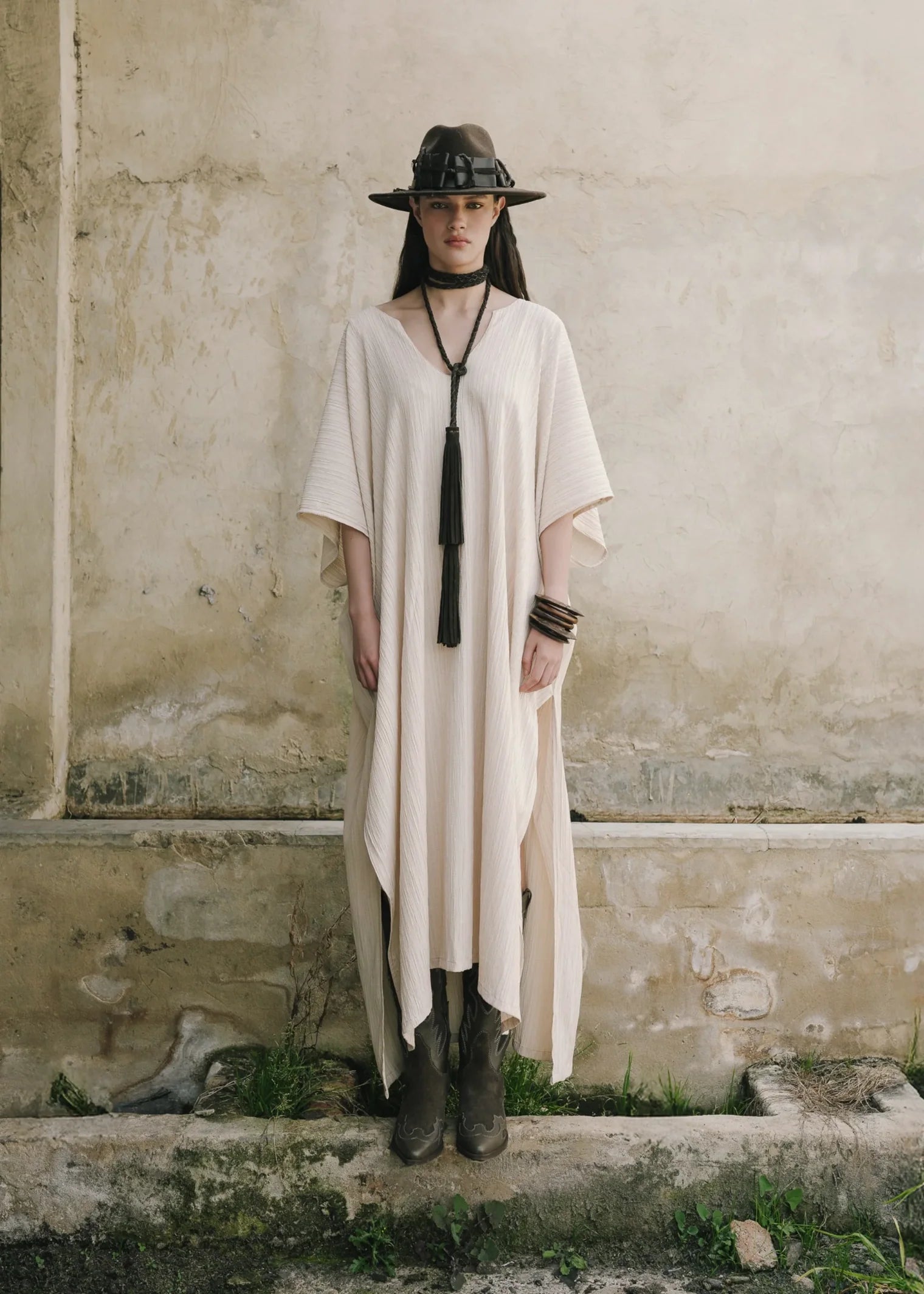 Person wearing a long beige dress with a hat and boots standing against a textured wall.