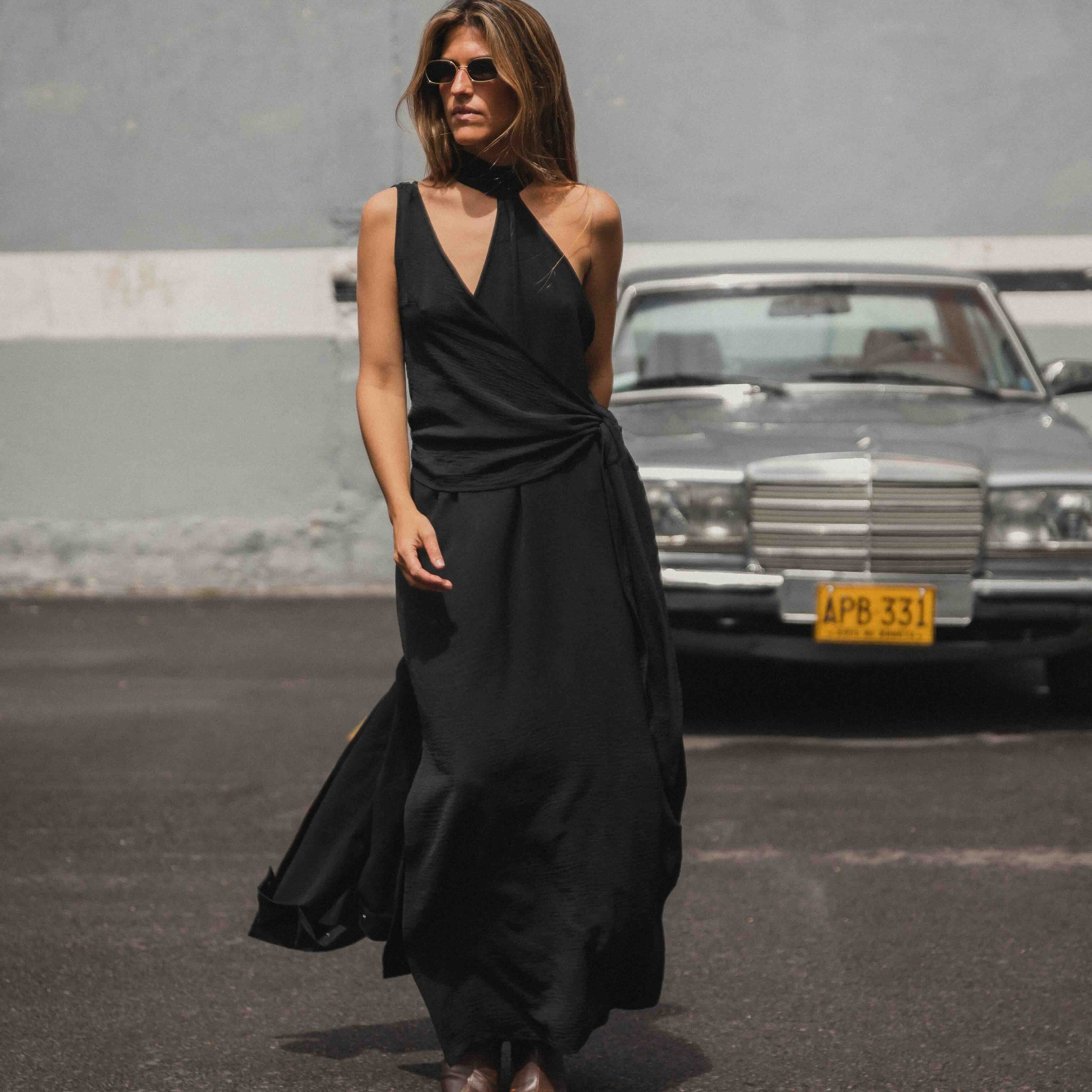 Woman in a black dress standing in an urban setting with a vintage car in the background.