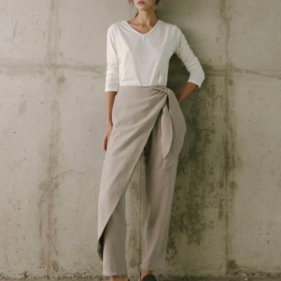 Woman wearing a white top and beige tied pants against a concrete wall.