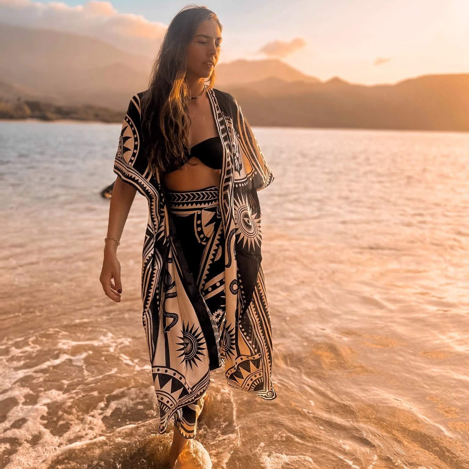 The designer of PLISSE, Paloma wearing the nazar kimono and wrap skirt. She is posing in the water at a beach during golden hour. This is the front of this flowy kimono.