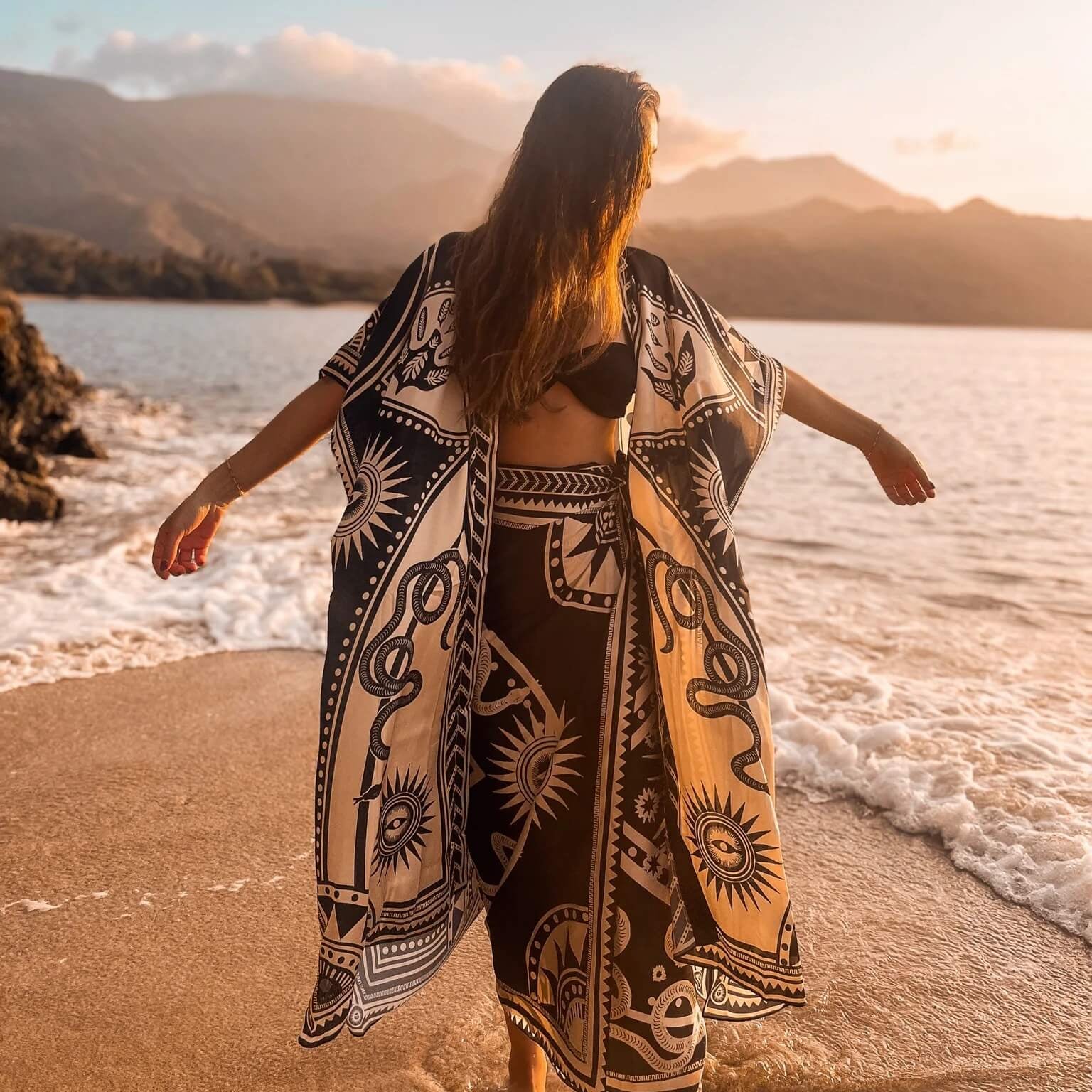 The designer of PLISSE, Paloma wearing the nazar kimono and wrap skirt. She is posing with her arms up on the sand at a beach during golden hour. This is the front of this flowy kimono.