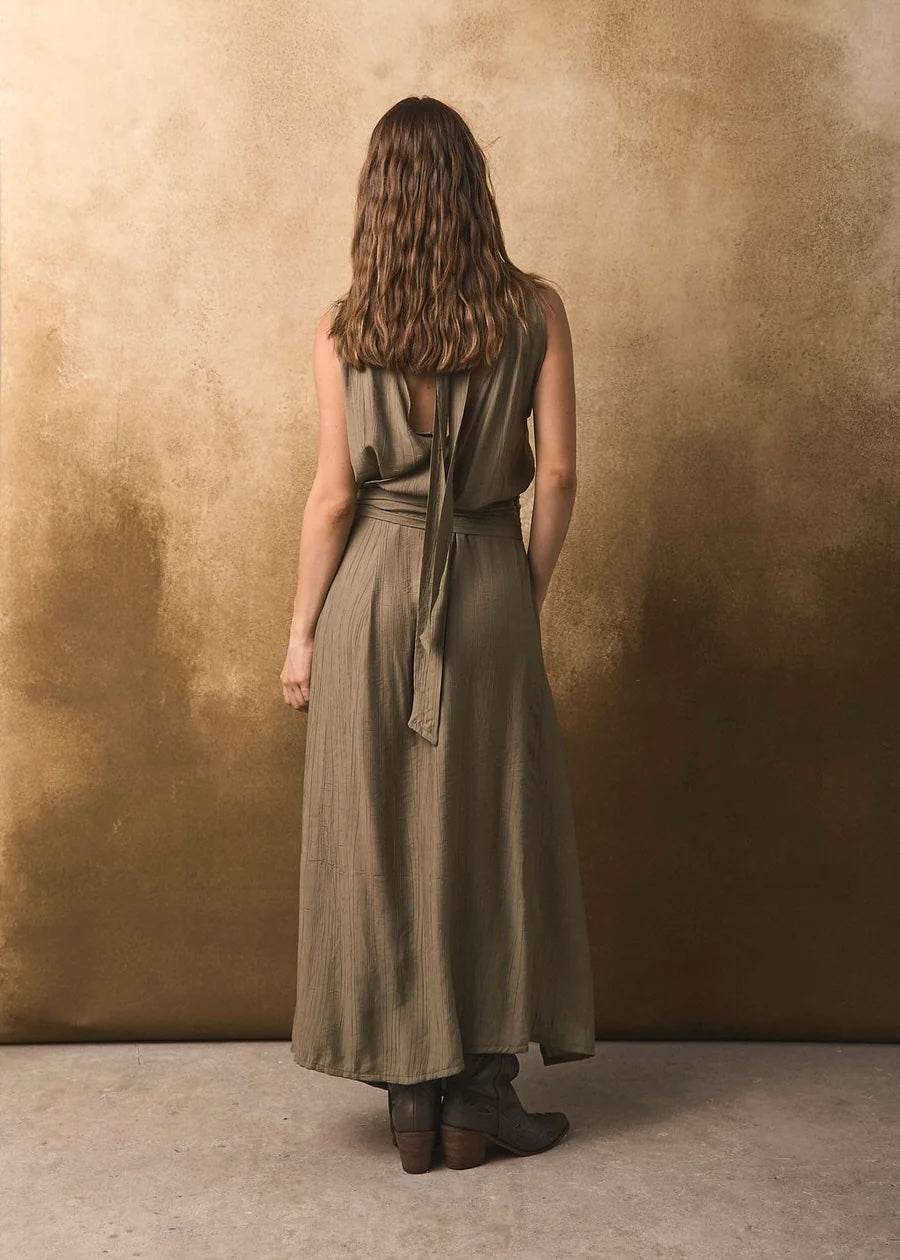 Back of a woman wearing a long olive green dress against a textured brown wall.