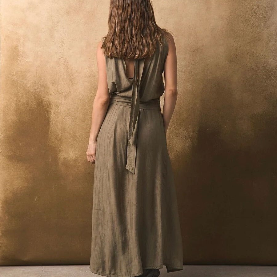 Back of a woman wearing a long olive green dress against a textured brown wall.