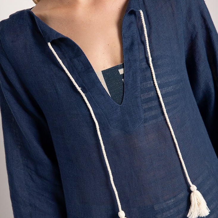 Close up image of a model wearing a blue cover-up with white cotton tassels on the neckline. She's standing against a white background. The purpose of this image is to highlight the cover-up.