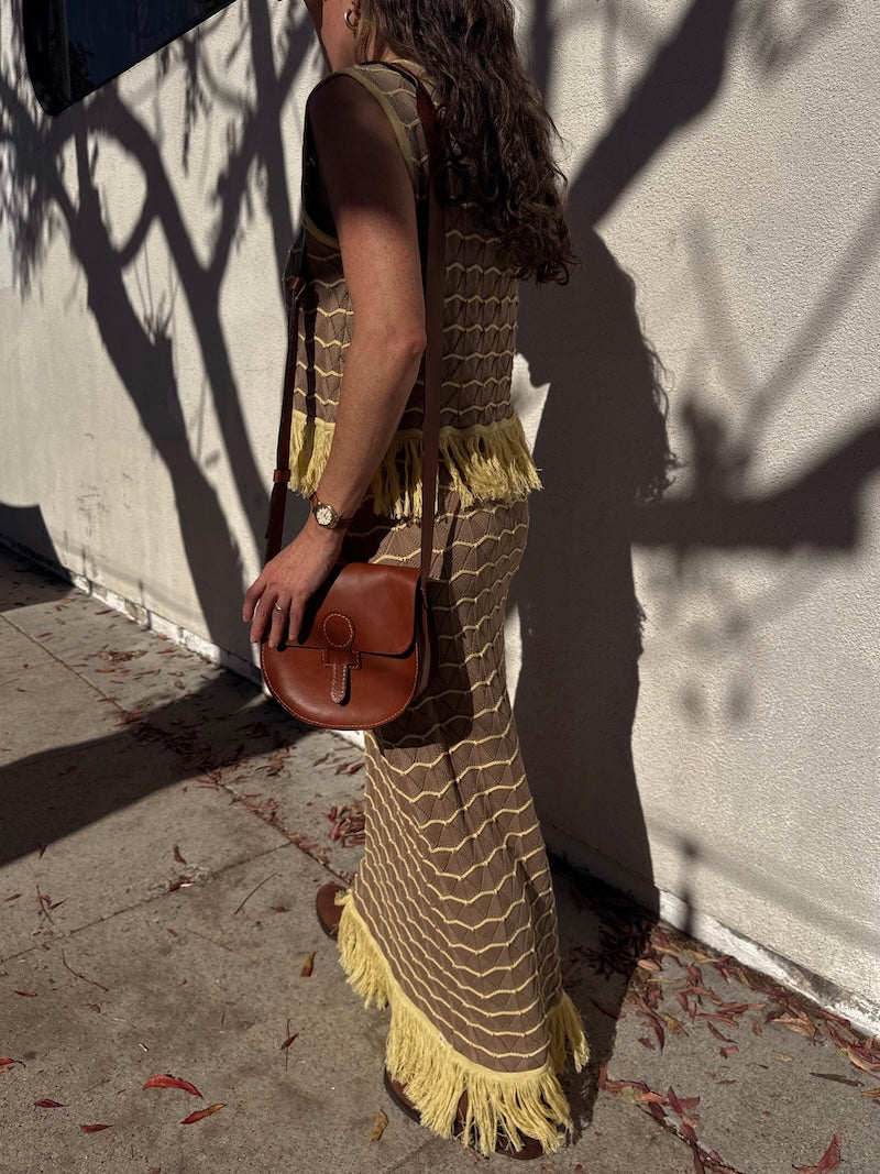 Woman in a long, patterned dress with yellow fringes holding a brown purse against a light-colored wall.