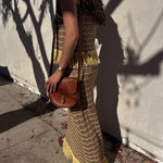 Woman in a long, patterned dress with yellow fringes holding a brown purse against a light-colored wall.