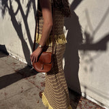 Woman in a long, patterned dress with yellow fringes holding a brown purse against a light-colored wall.