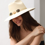 Woman wearing a beige hat with a wide brim and decorative crystals, sitting against a plain background.