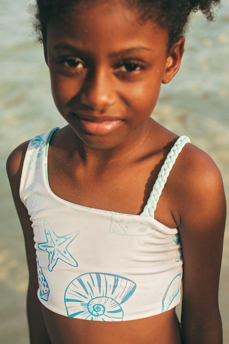 Young girl wearing a white tank top with blue designs on a beach.