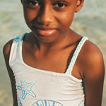Young girl wearing a white tank top with blue designs on a beach.