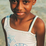 Young girl wearing a white tank top with blue designs on a beach.