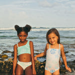 Two young girls in swimsuits standing in shallow water at the beach.