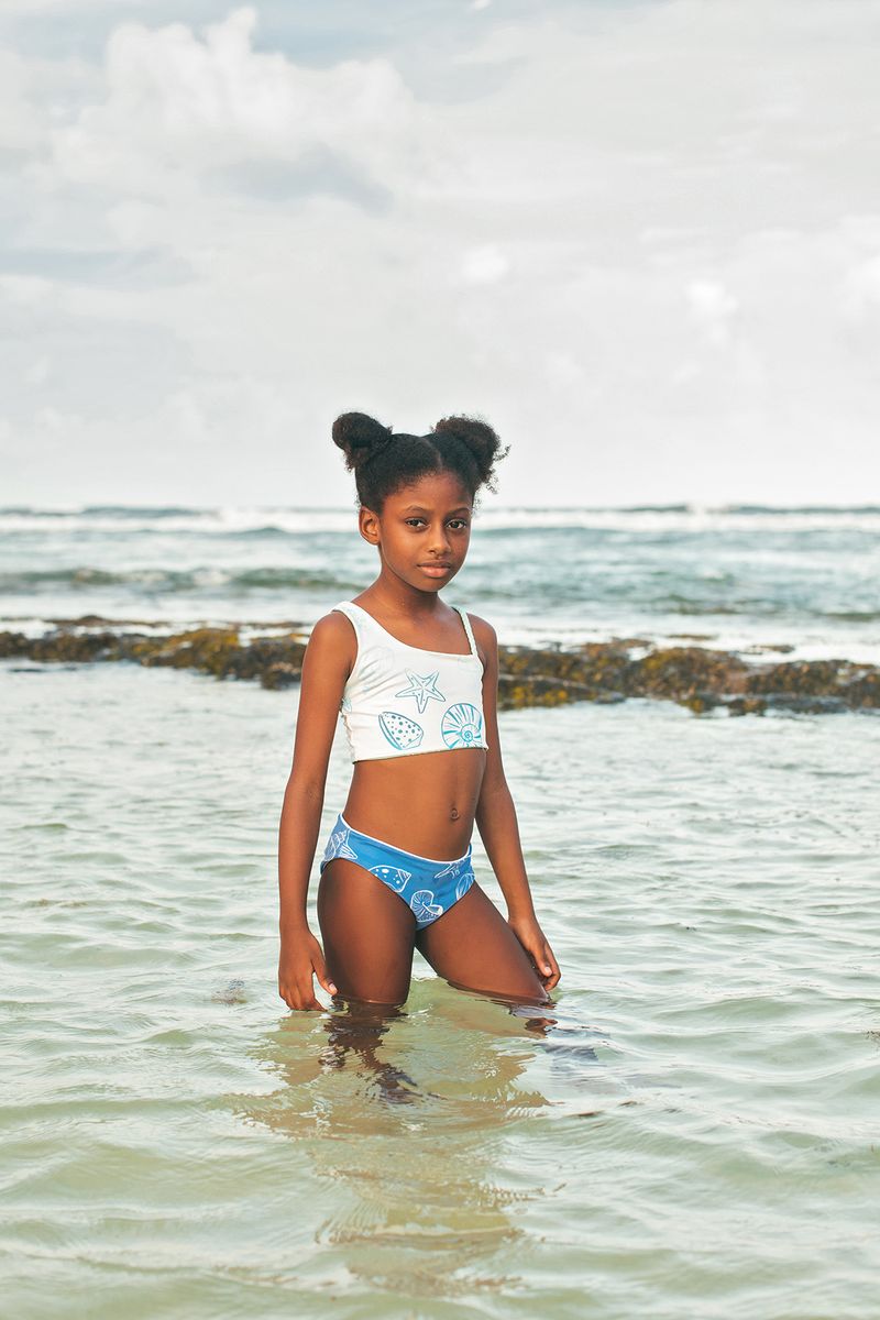 Young girl in a white tank top and blue shorts standing in shallow water at the beach.