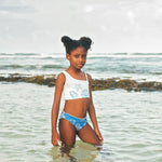 Young girl in a white tank top and blue shorts standing in shallow water at the beach.