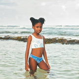 Young girl in a white tank top and blue shorts standing in shallow water at the beach.