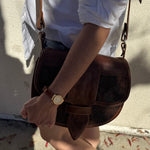Person holding a brown leather bag with a gold watch on wrist, wearing a white shirt and denim shorts.