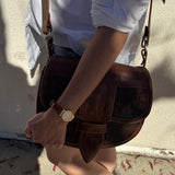 Person holding a brown leather bag with a gold watch on wrist, wearing a white shirt and denim shorts.