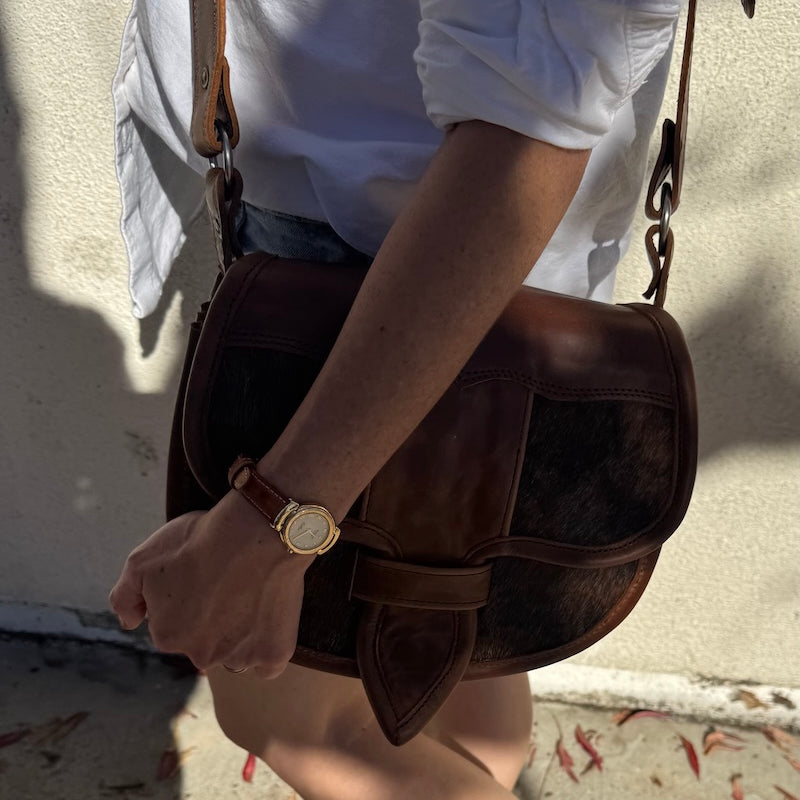 Person holding a brown leather bag with a gold watch on wrist, wearing a white shirt and denim shorts.