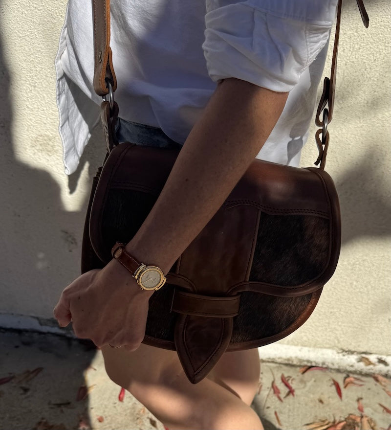 Person holding a brown leather bag with a gold watch on wrist, wearing a white shirt and denim shorts.