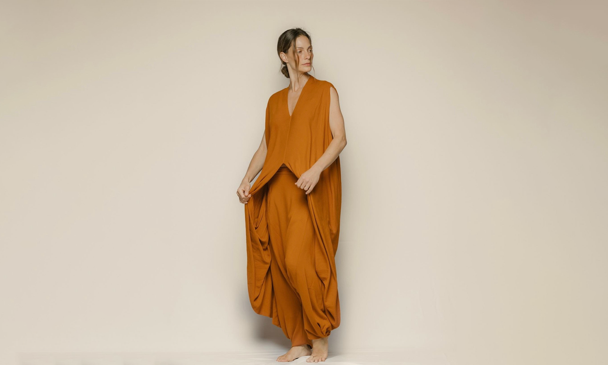 Image of a woman wearing a terracotta jumpsuit. She's standing against a light background and looking to the side.