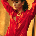 Image of a woman wearing gold earrings and a matching pin. She's also wearing a red dress and seems to be getting ready, putting her hair back. The purpose of this image is to highlight the earrings by Alma jewelry.