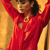Image of a woman wearing gold earrings and a matching pin. She's also wearing a red dress and seems to be getting ready, putting her hair back. The purpose of this image is to highlight the earrings by Alma jewelry.