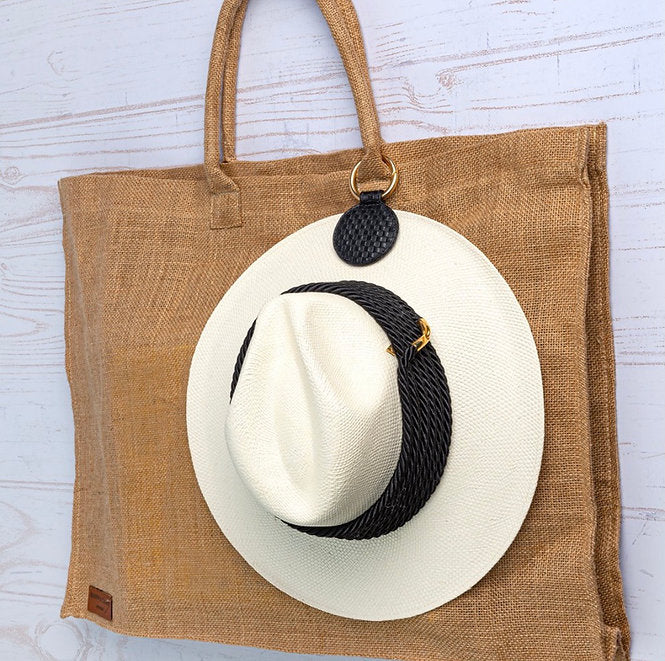 White straw hat with black band attached to a black brown tote bag using black pendant against a light wooden background