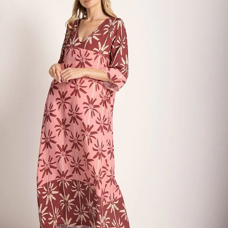 Woman wearing a long cover-up dress with a pink and tan palm tree print. She's looking towards the camera and standing in front of a white background. The purpose of this image is to highlight the dress.