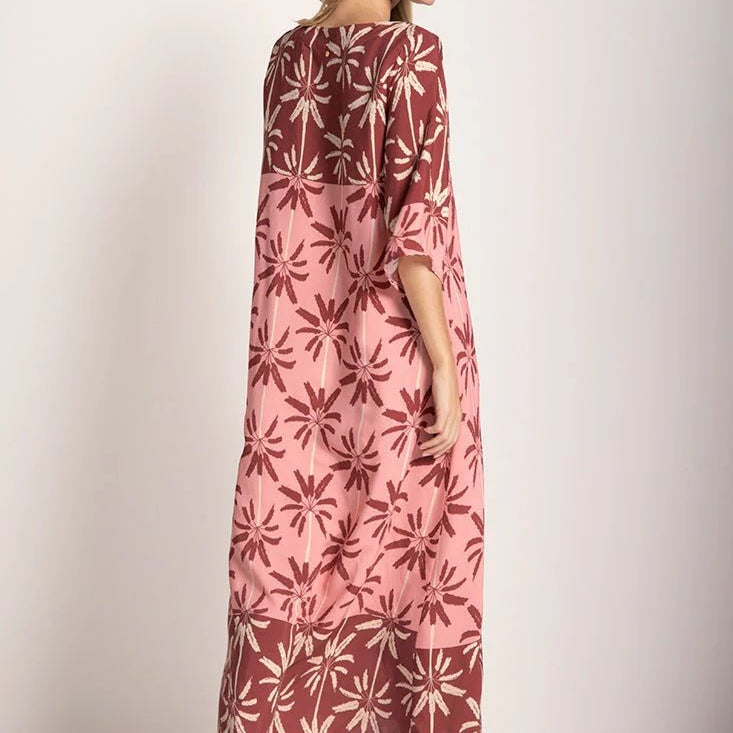 Woman wearing a long cover-up dress with a pink and tan palm tree print. She's looking towards the side and giving her back to the camera. She's standing in front of a white background. The purpose of this image is to highlight the dress.