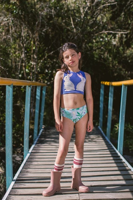 Girl in a two-piece bathing suit set with a blue bikini top and patterned bikini bottoms standing on a wooden bridge with greenery in the background.