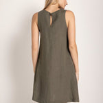 Image of a woman wearing a short linen dress that is brown/grey in color. She's standing against a white background and looking to the side. We're able to see her back. The purpose of this image is to highlight the dress.