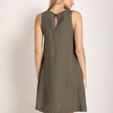 Image of a woman wearing a short linen dress that is brown/grey in color. She's standing against a white background and looking to the side. We're able to see her back. The purpose of this image is to highlight the dress.