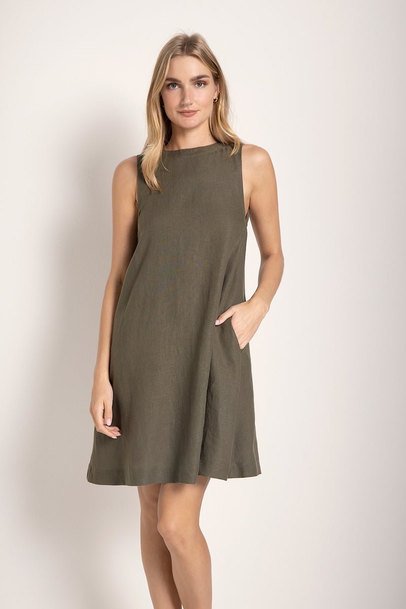 Image of a woman wearing a short linen dress that is brown/grey in color. She's standing against a white background and has one hand on her pocket. The purpose of this image is to highlight the dress.
