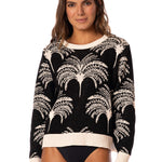 Woman wearing a sweater with blakc and white palm tree pattern and black swim bottoms on a white background.
