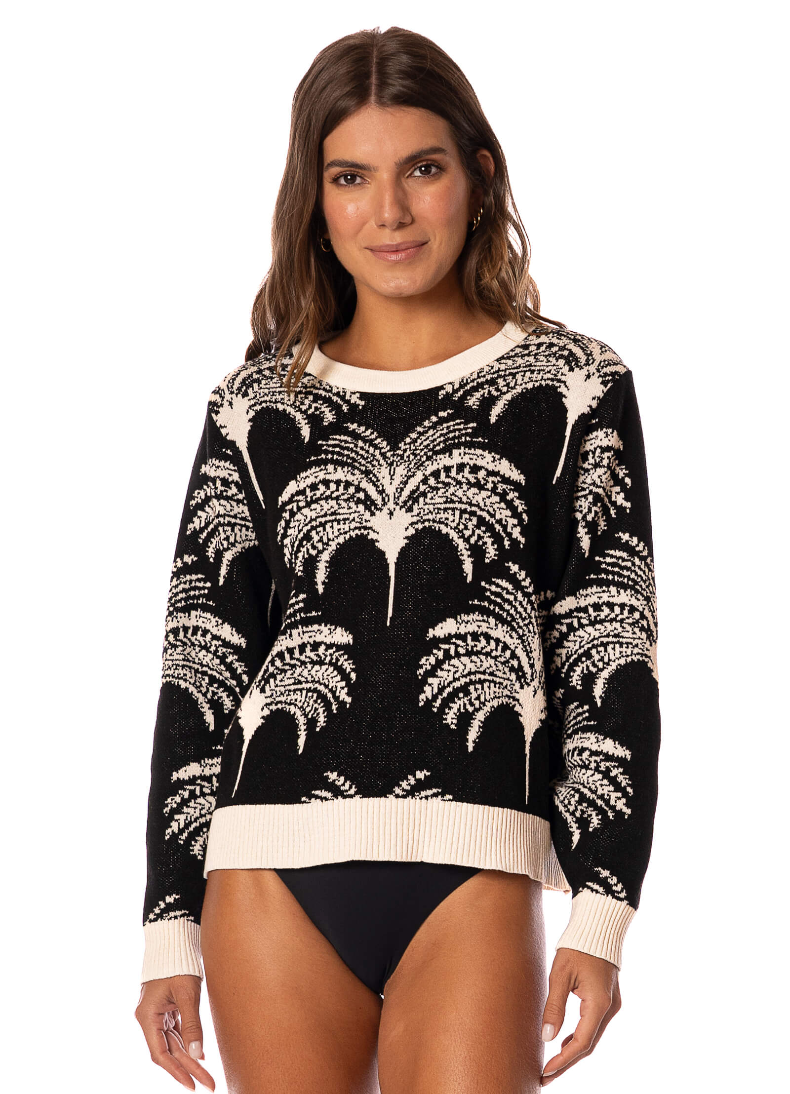 Woman wearing a sweater with blakc and white palm tree pattern and black swim bottoms on a white background.