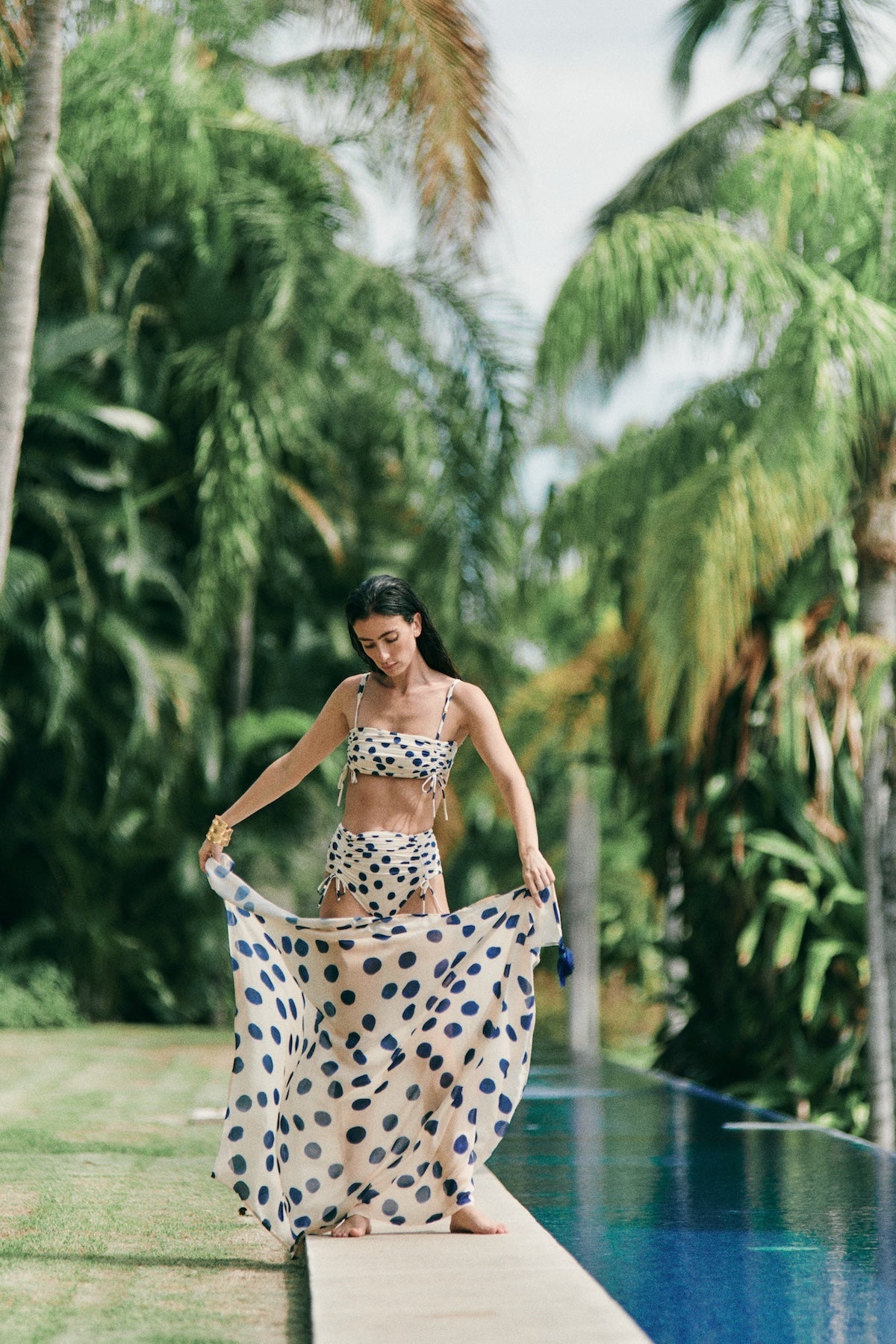 Woman wearing a white swimsuit with blue polka dots. She's holding a matching sarong and standing along the edge of a pool. The purpose of this image is to highlight the swimsuit and sarong because they're for sale at EVAMAIA Boutique.