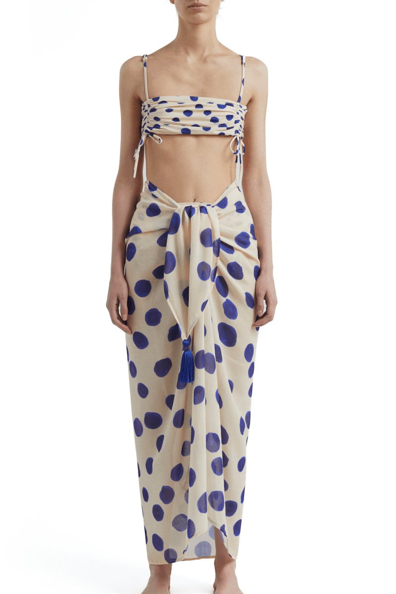 Woman wearing a bikini top with blue polka dots and a matching sarong. She's standing behind a white background. The purpose of this image is to highlight the sarong by Encantadore Swimwear.