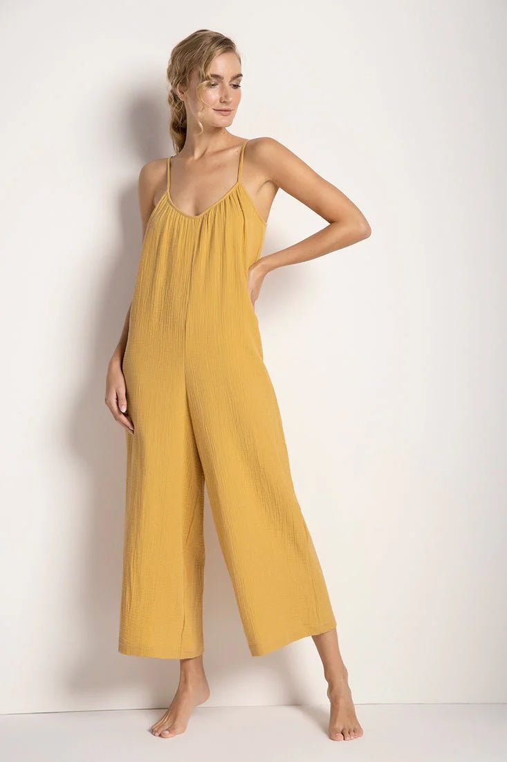 Woman wearing a rusted yellow jumpsuit with tie detail on the sleeves. She's standing in front of a white background and looking to the side. The purpose of this image is to highlight the jumpsuit.