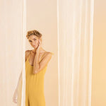 Woman wearing a rusted yellow jumpsuit with tie detail on the sleeves. She's standing in front of a white curtain in the background and looking to the camera. The purpose of this image is to highlight the jumpsuit.