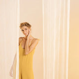 Woman wearing a rusted yellow jumpsuit with tie detail on the sleeves. She's standing in front of a white curtain in the background and looking to the camera. The purpose of this image is to highlight the jumpsuit.