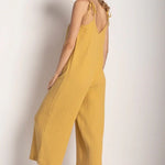 Woman wearing a rusted yellow jumpsuit with tie detail on the sleeves. She's standing in front of a white background and looking to the camera while showing the back of the jumpsuit. The purpose of this image is to highlight the jumpsuit.