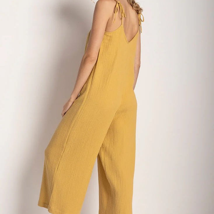 Woman wearing a rusted yellow jumpsuit with tie detail on the sleeves. She's standing in front of a white background and looking to the camera while showing the back of the jumpsuit. The purpose of this image is to highlight the jumpsuit.