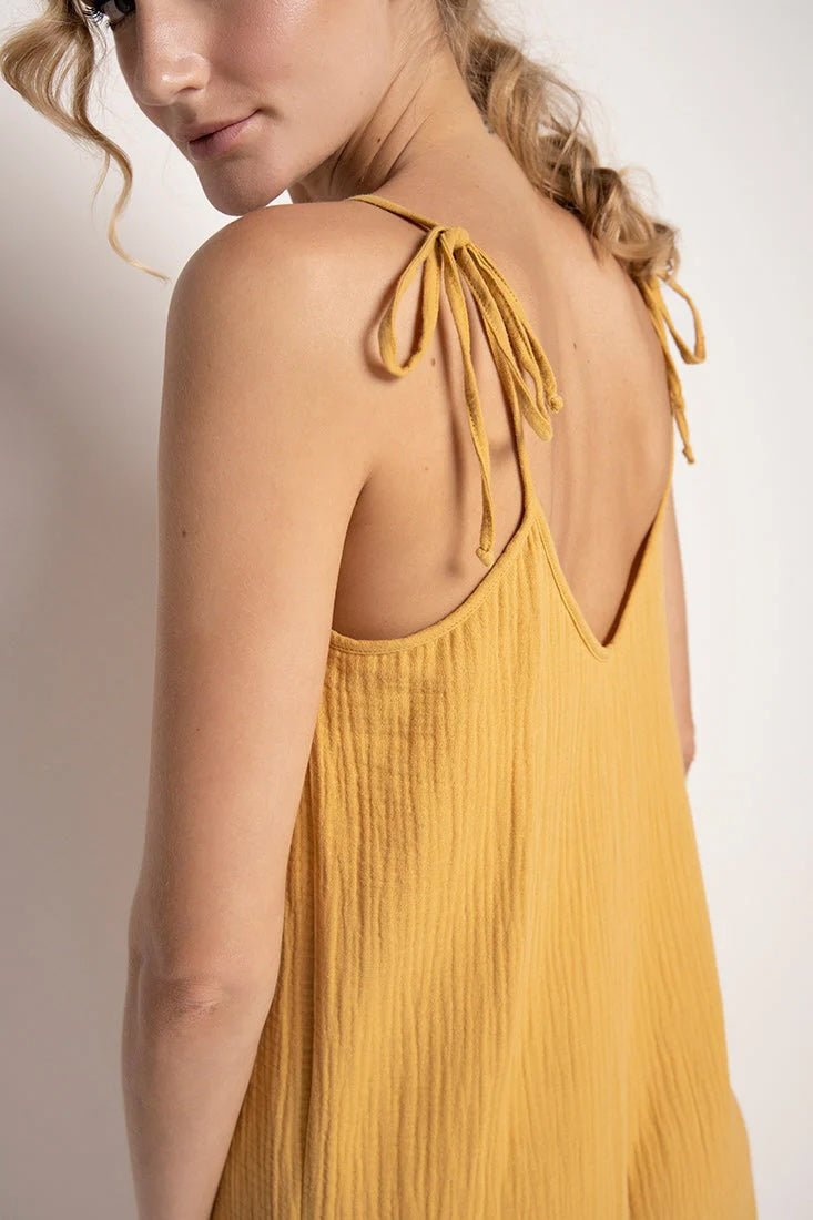 Close up of a woman wearing a rusted yellow jumpsuit with tie detail on the sleeves. She's standing in front of a white background and looking to the camera. The purpose of this image is to highlight the tie sleeves on the jumpsuit.