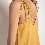 Close up of a woman wearing a rusted yellow jumpsuit with tie detail on the sleeves. She's standing in front of a white background and looking to the camera. The purpose of this image is to highlight the tie sleeves on the jumpsuit.