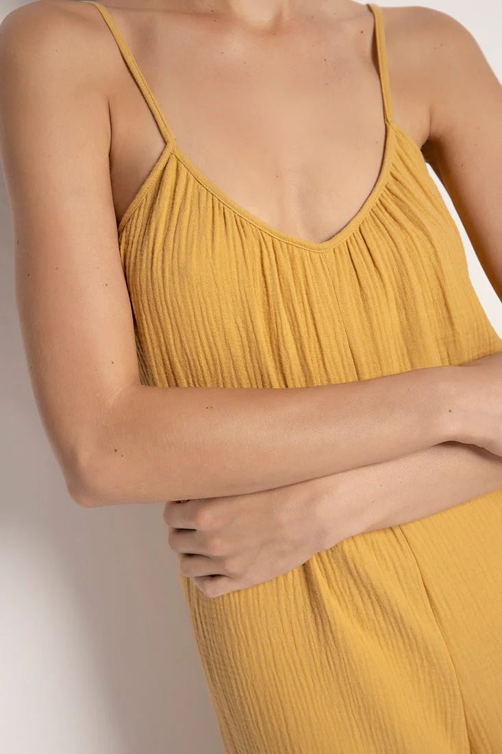 Close up image of a woman wearing a rusted yellow jumpsuit with tie detail on the sleeves. She's standing in front of a white background. The purpose of this image is to highlight the neckline of the jumpsuit.