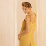Woman wearing a rusted yellow jumpsuit with tie detail on the sleeves. She's standing in front of a white curtain and looking to the side. The purpose of this image is to highlight the jumpsuit.