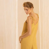 Woman wearing a rusted yellow jumpsuit with tie detail on the sleeves. She's standing in front of a white curtain and looking to the side. The purpose of this image is to highlight the jumpsuit.