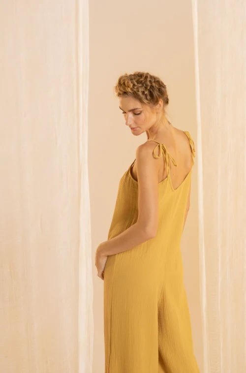 Woman wearing a rusted yellow jumpsuit with tie detail on the sleeves. She's standing in front of a white curtain and looking to the side. The purpose of this image is to highlight the jumpsuit.