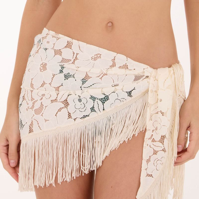 An off-white lace wrap sarong with floral embroidery and long fringe, worn as a hip scarf by a model.