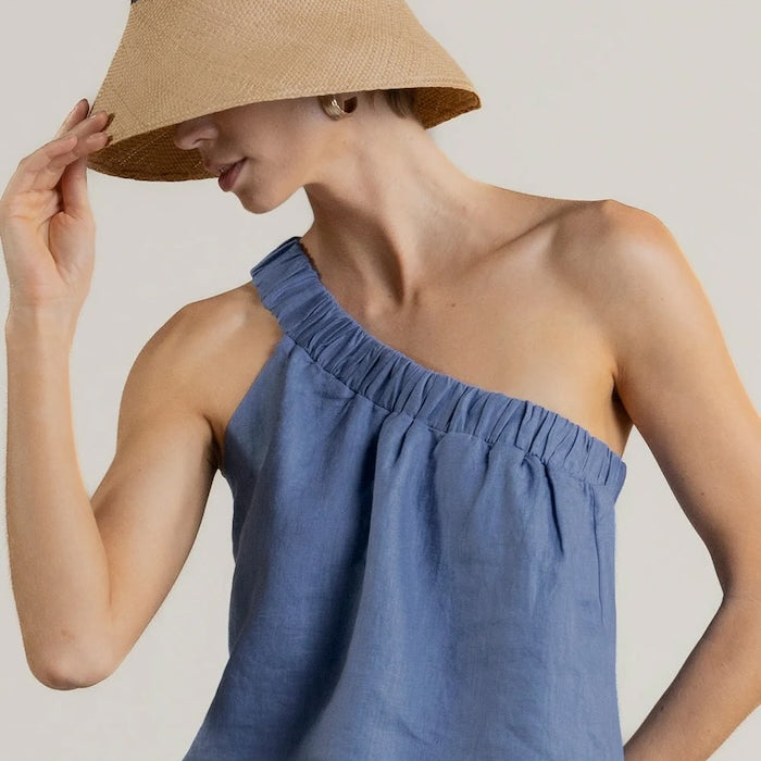 Woman wearing a blue one-shoulder top and beige bucket hat on a plain background.